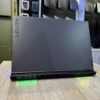 A Lenovo Legion gaming laptop showcased on a wooden table with visible green LED lighting and various ports, surrounded by blurred shelves of other laptops and products.