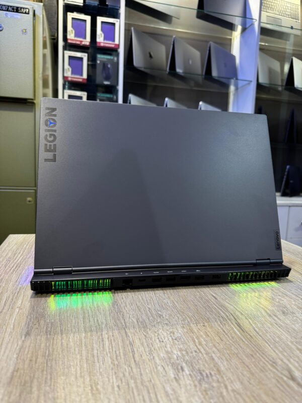 A Lenovo Legion gaming laptop showcased on a wooden table with visible green LED lighting and various ports, surrounded by blurred shelves of other laptops and products.